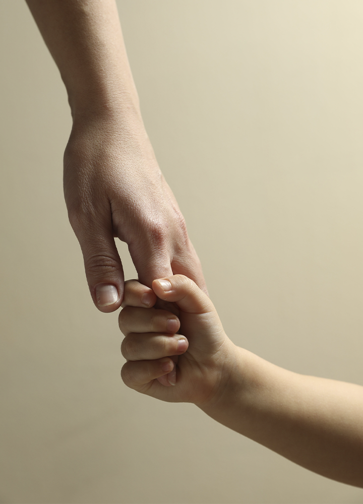 Mano de una persona adulta sujetando la mano de una niña o niño pequeño sobre un fondo liso y desenfocado. La imagen se centra en el gesto de apoyo y acompañamiento entre ambas.