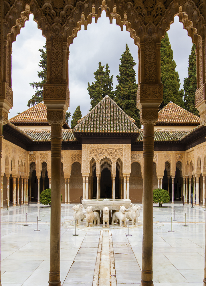 Patio histórico con arcos y columnas ornamentadas que enmarcan una fuente de mármol en el centro. La arquitectura decorada y los tejados de teja destacan sobre un fondo de árboles altos.