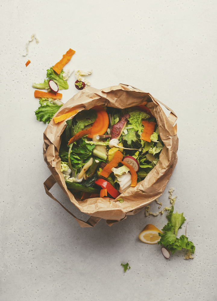 Bolsa de papel abierta con restos de una ensalada colorida compuesta por hojas verdes, zanahoria, rábano y otras verduras. Alrededor aparecen pequeños fragmentos de alimentos sobre una superficie clara.