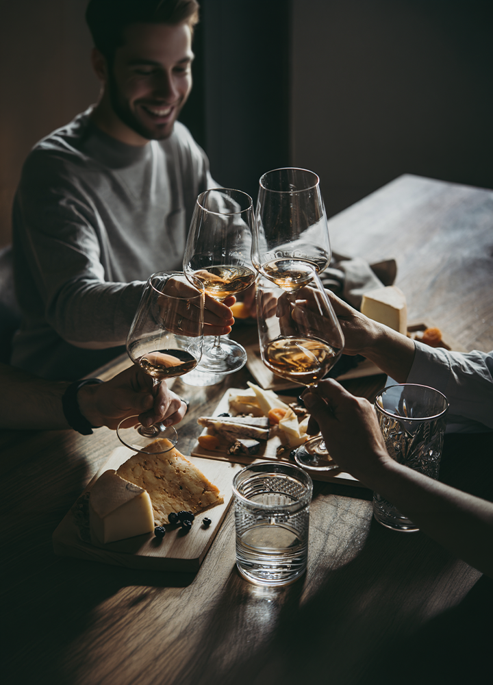 Varias personas brindando con copas de vino alrededor de una mesa de madera con una selección de quesos y aperitivos. La escena está iluminada con luz tenue y cálida, destacando las copas en el centro de la composición. Sobre la mesa se ven tablas de quesos, vasos de agua y elementos de servicio, mientras las personas se reúnen alrededor compartiendo una experiencia gastronómica.