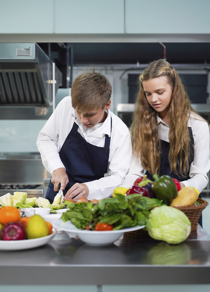 Dos personas con delantal trabajan en una cocina profesional preparando alimentos sobre una mesa de acero inoxidable. En primer plano se observan verduras y frutas variadas, como lechuga, col, pimientos, tomates, cebolla, pera y hojas verdes, mientras una de las personas corta ingredientes sobre una tabla y la otra colabora en la preparación. Al fondo se distinguen equipos de cocina industrial y superficies metálicas propias de un entorno de formación o trabajo culinario.