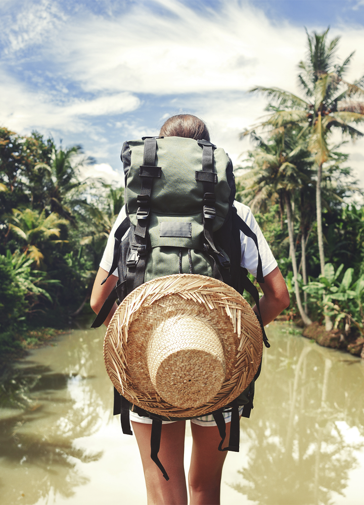 Persona de espaldas observa un paisaje tropical junto a un río o canal rodeado de abundante vegetación y palmeras. Lleva una mochila de senderismo de gran tamaño con un sombrero de paja sujeto en la parte trasera, mientras el agua refleja parcialmente los árboles bajo un cielo con nubes dispersas.