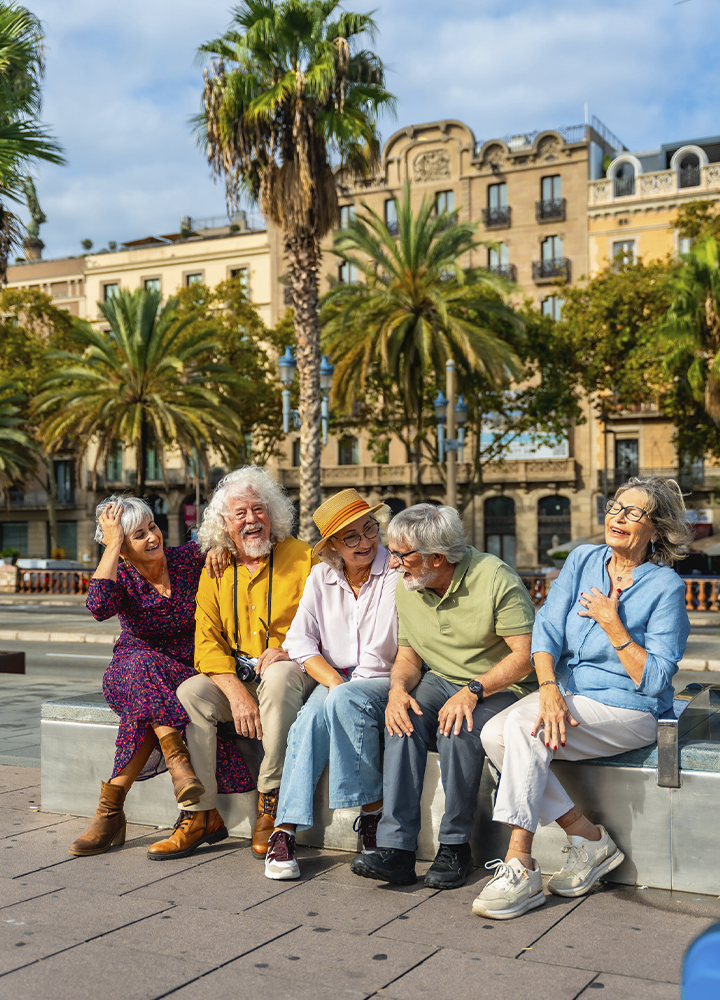 Cinco personas mayores sentadas en un banco de una plaza urbana rodeada de palmeras y edificios históricos. El grupo conversa mientras descansa al aire libre en un entorno soleado.