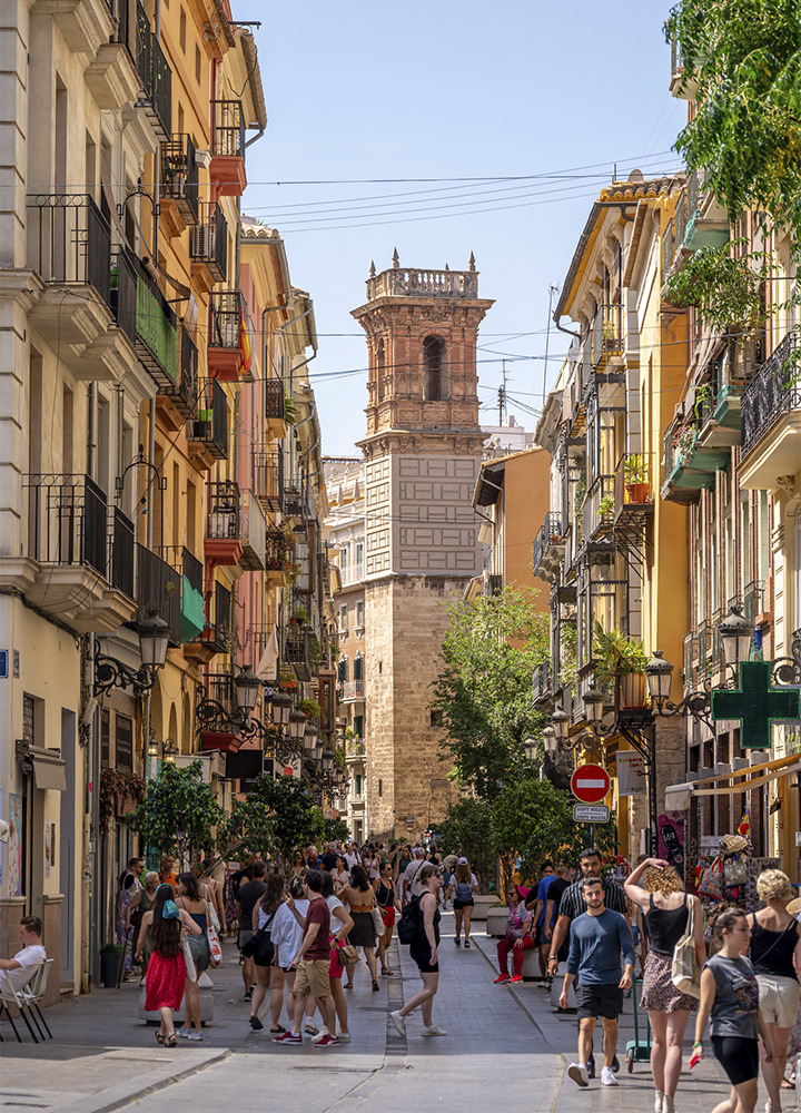 Calle peatonal concurrida flanqueada por edificios históricos con balcones de forja y fachadas en tonos cálidos. Al fondo se eleva una torre de piedra de estilo histórico, visible entre las construcciones. Numerosas personas pasean por la calle, que está decorada con árboles y comercios a ambos lados.