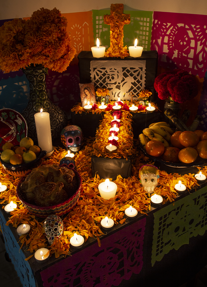 Altar tradicional decorado con flores, frutas, velas encendidas y calaveras ornamentales. La composición, iluminada con luz cálida, destaca una cruz central rodeada de elementos simbólicos y papel decorativo de colores.