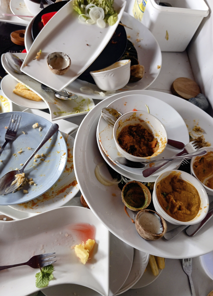 Montón de platos, cuencos y cubiertos usados con restos de comida sobre una mesa. Se observan migas, salsas y recipientes vacíos apilados, reflejando desperdicio alimentario tras una comida.
