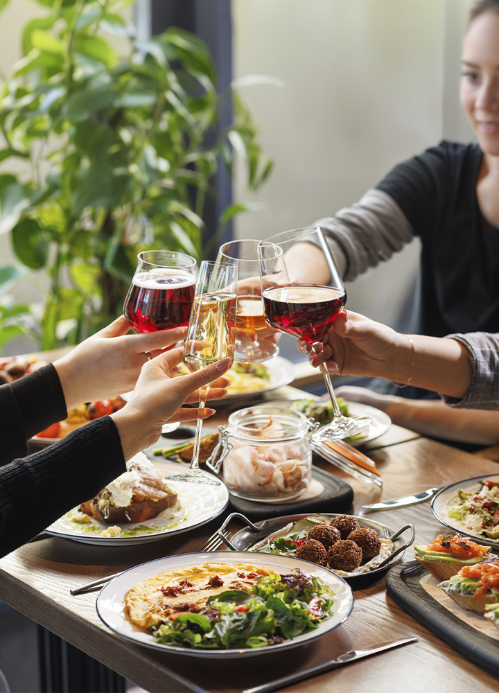 Varias personas brindando con copas de vino y cava alrededor de una mesa con distintos platos para compartir. Sobre la mesa se observan ensaladas, tostadas, aperitivos, cubiertos y un tarro de cristal en el centro. Al fondo hay una planta de interior junto a una ventana que aporta luz natural a la escena, creando un ambiente de reunión gastronómica en un espacio acogedor.