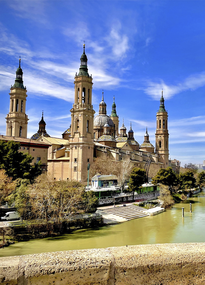 Vista de la Basílica del Pilar junto al río Ebro en Zaragoza. El edificio destaca por sus cuatro torres, varias cúpulas decoradas y una gran cúpula central, elevándose sobre la ribera arbolada. En primer plano se observa el cauce del río de color verdoso y parte de un puente de piedra. El cielo azul con nubes alargadas aporta luminosidad a la escena y resalta la silueta monumental de la basílica.