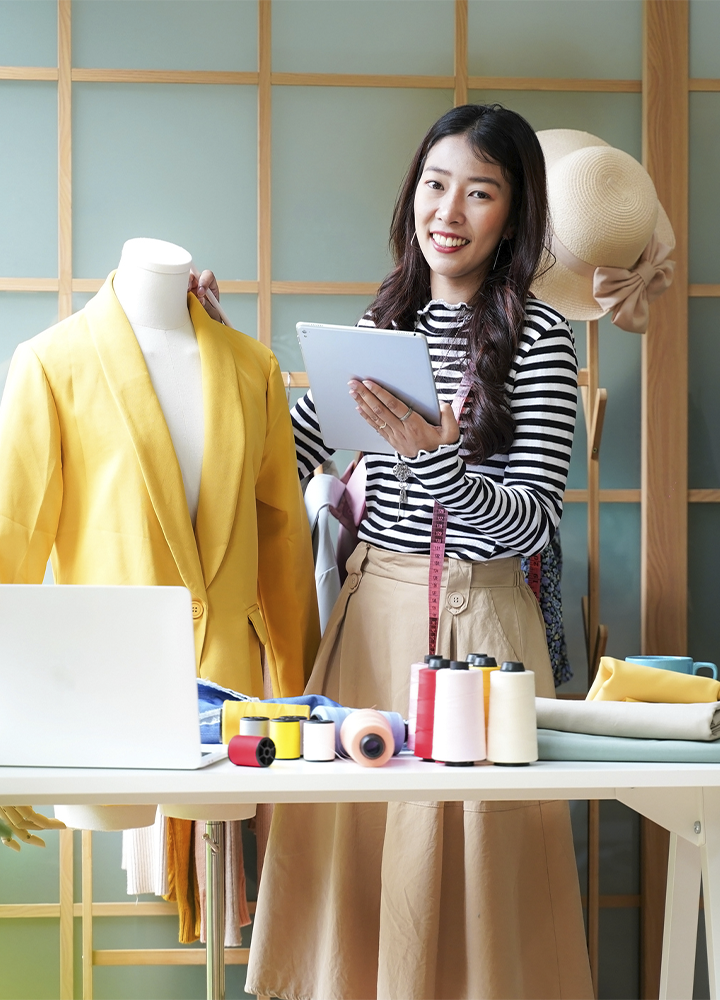 Persona sostiene una tableta digital junto a un maniquí vestido con una chaqueta amarilla en un estudio de diseño de moda. Sobre la mesa hay carretes de hilo de varios colores, tejidos, una cinta métrica y un ordenador portátil, mientras al fondo se aprecia un perchero con prendas y un sombrero colgado.