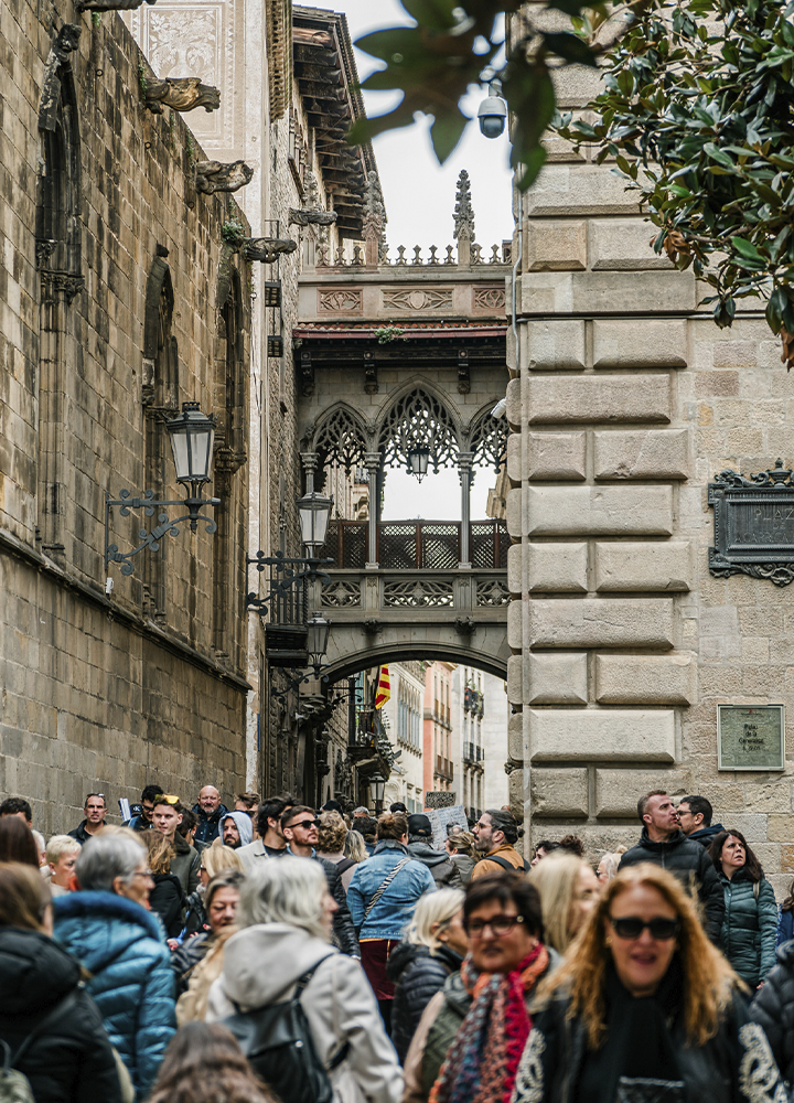 Calle histórica muy concurrida, flanqueada por edificios de piedra, con un pasadizo elevado de estilo gótico que conecta dos construcciones sobre la vía. Numerosos peatones recorren el estrecho corredor urbano mientras faroles y elementos ornamentales decoran las fachadas.