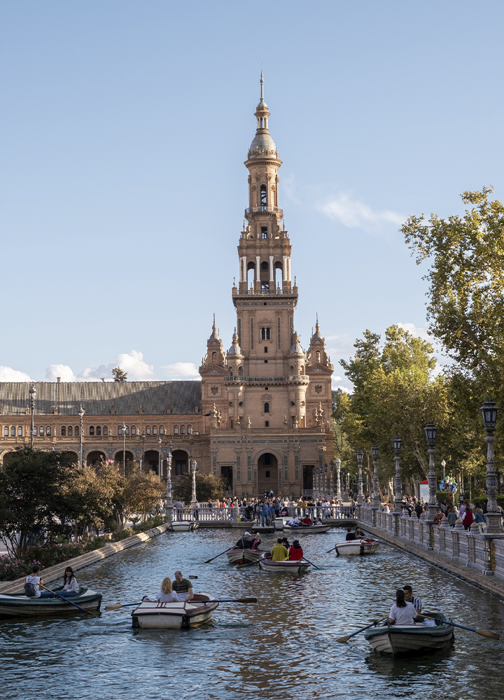 Canal de agua frente a un gran edificio histórico con una alta torre central. Varias barcas navegan por el canal mientras numerosas personas pasean por las orillas. Árboles y farolas bordean el recorrido bajo un cielo azul despejado.