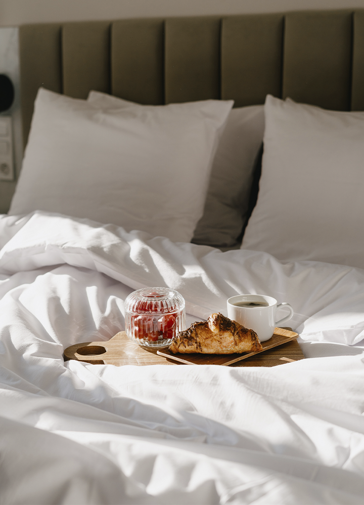 Bandeja de desayuno sobre una cama con sábanas blancas deshechas. La bandeja de madera contiene un cruasán dorado, una taza de café y un pequeño recipiente de cristal con mermelada roja. Al fondo, dos almohadas blancas apoyadas en un cabecero acolchado de color oscuro. La luz natural entra lateralmente y crea sombras suaves sobre la ropa de cama.