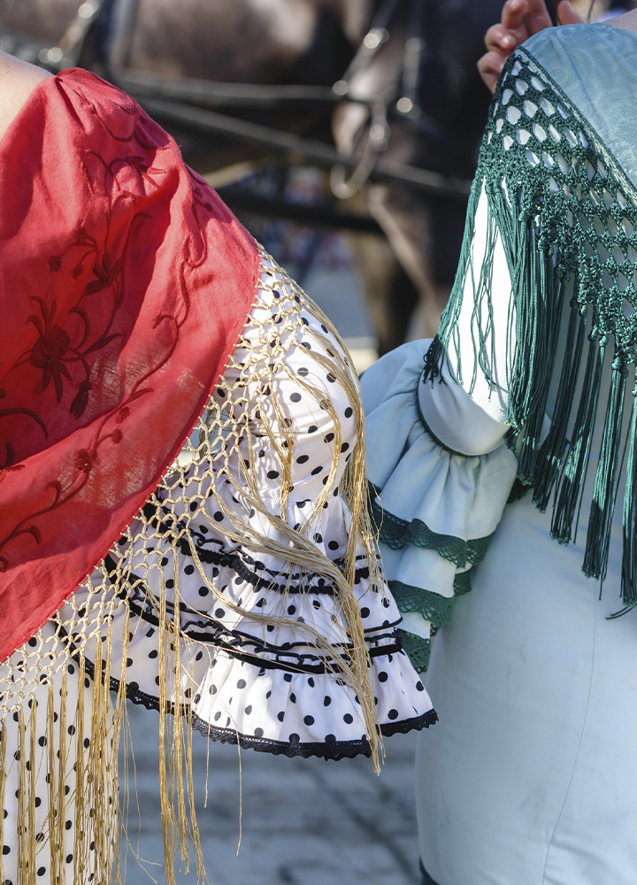 Detalle de dos trajes tradicionales con mantones de flecos, uno rojo bordado y otro verde, vistos de cerca. Los tejidos, encajes y adornos ocupan casi toda la imagen y destacan por sus colores y texturas.