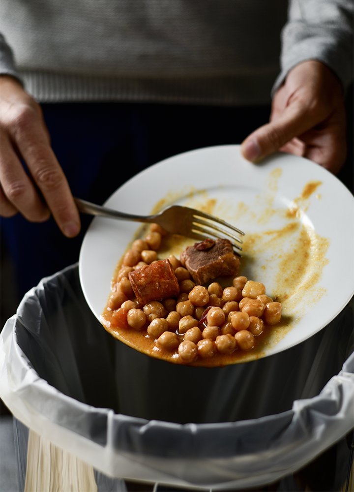 Plato con garbanzos y trozos de comida sostenido sobre un cubo de basura. Una mano sujeta un tenedor mientras los restos están a punto de desecharse, ilustrando el desperdicio alimentario.