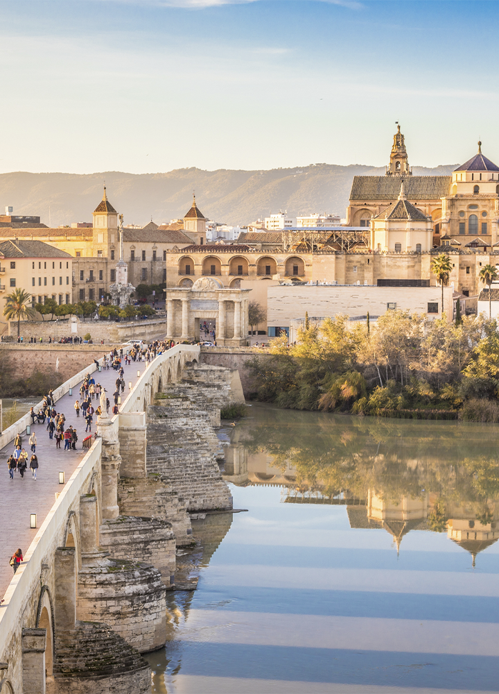 Vista de la Mezquita-Catedral de Córdoba y su entorno histórico junto al río Guadalquivir. En primer plano destaca el Puente Romano, recorrido por numerosas personas, mientras los edificios monumentales se reflejan en las aguas tranquilas del río. Al fondo se aprecian las montañas bajo una luz cálida de final de día.