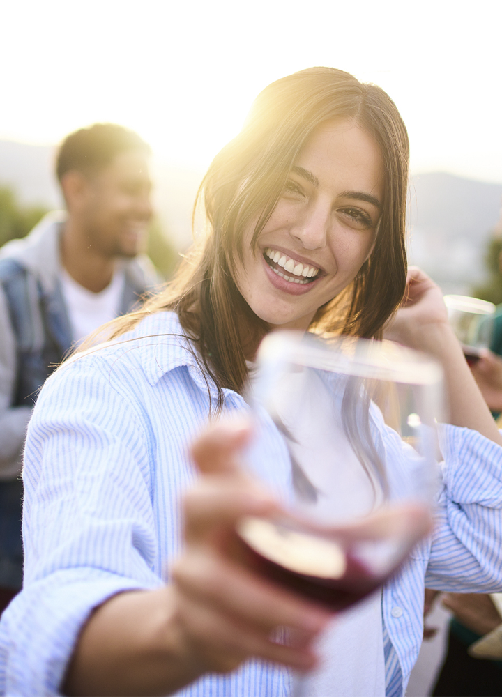 Persona en primer plano sosteniendo una copa de vino tinto hacia la cámara en una reunión al aire libre. La escena está iluminada por la luz del sol a contraluz, que realza el contorno de la figura. Al fondo aparecen otras personas desenfocadas con copas en la mano y un paisaje exterior suavemente difuminado. La copa ocupa el primer plano de la composición y aporta profundidad a la imagen.
