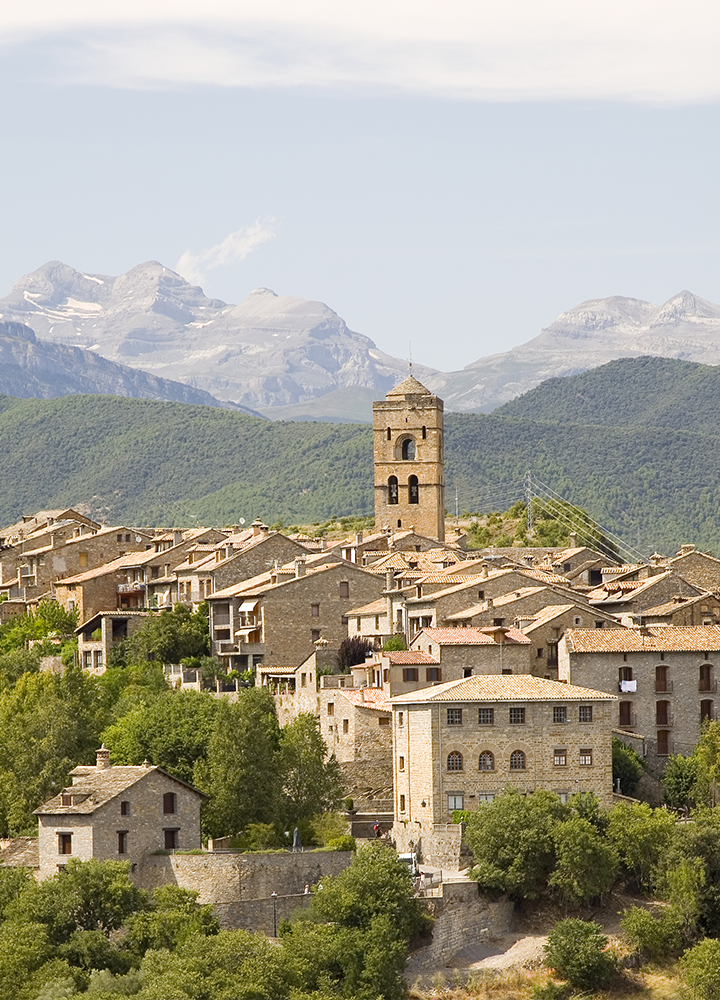 Vista de un pueblo de montaña con casas de piedra y tejados de teja agrupadas en torno a una torre campanario situada en el centro. La localidad está rodeada de colinas cubiertas de vegetación y, al fondo, se alzan montañas de cumbres rocosas con restos de nieve. La luz suave resalta los tonos cálidos de los edificios y el entorno natural que rodea el paisaje.