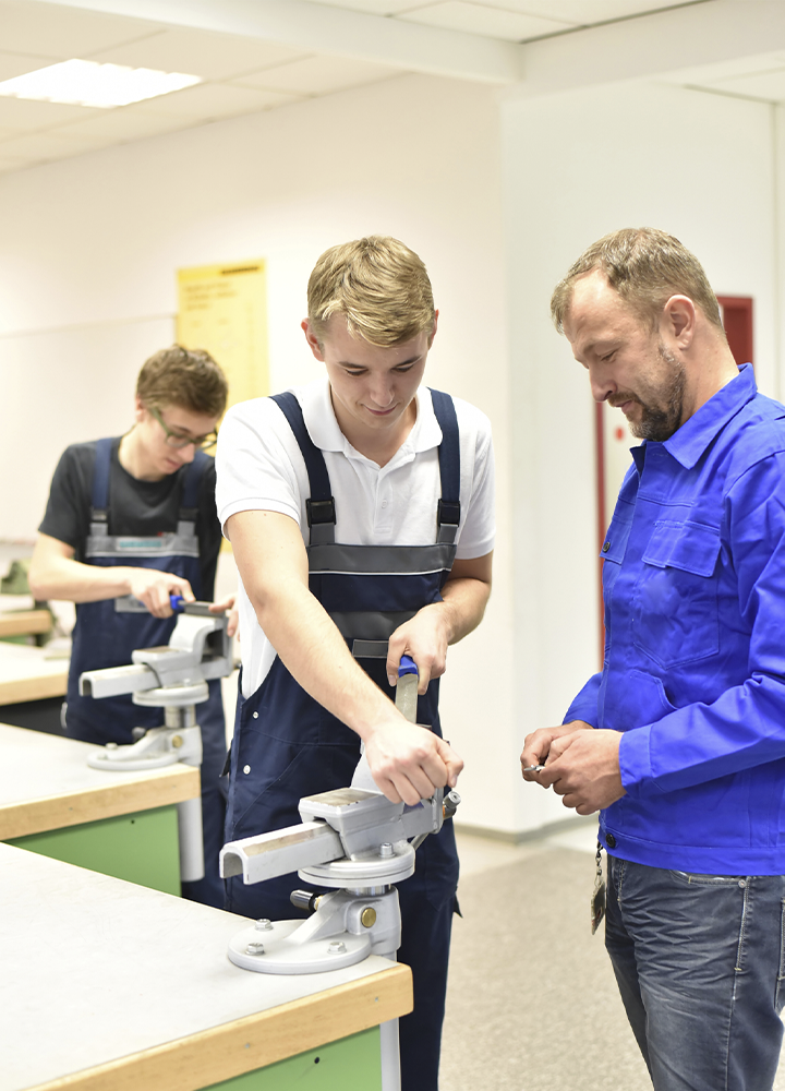 Dos personas y un instructor trabajan con tornillos de banco en un taller de formación técnica. En primer plano, una persona con peto de trabajo ajusta una pieza mientras otra con chaqueta azul observa la práctica. Al fondo, una tercera persona realiza una tarea similar en otra mesa de trabajo, en un aula equipada para actividades de aprendizaje práctico.