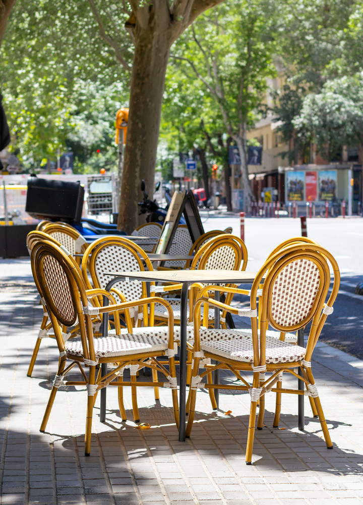 Terraza de cafetería situada en una acera arbolada, con varias mesas cuadradas rodeadas de sillas de mimbre en tonos amarillos y blancos. Al fondo se aprecia una calle urbana con vehículos, señalización y edificios parcialmente ocultos por la vegetación, mientras la luz del sol proyecta sombras sobre el pavimento.