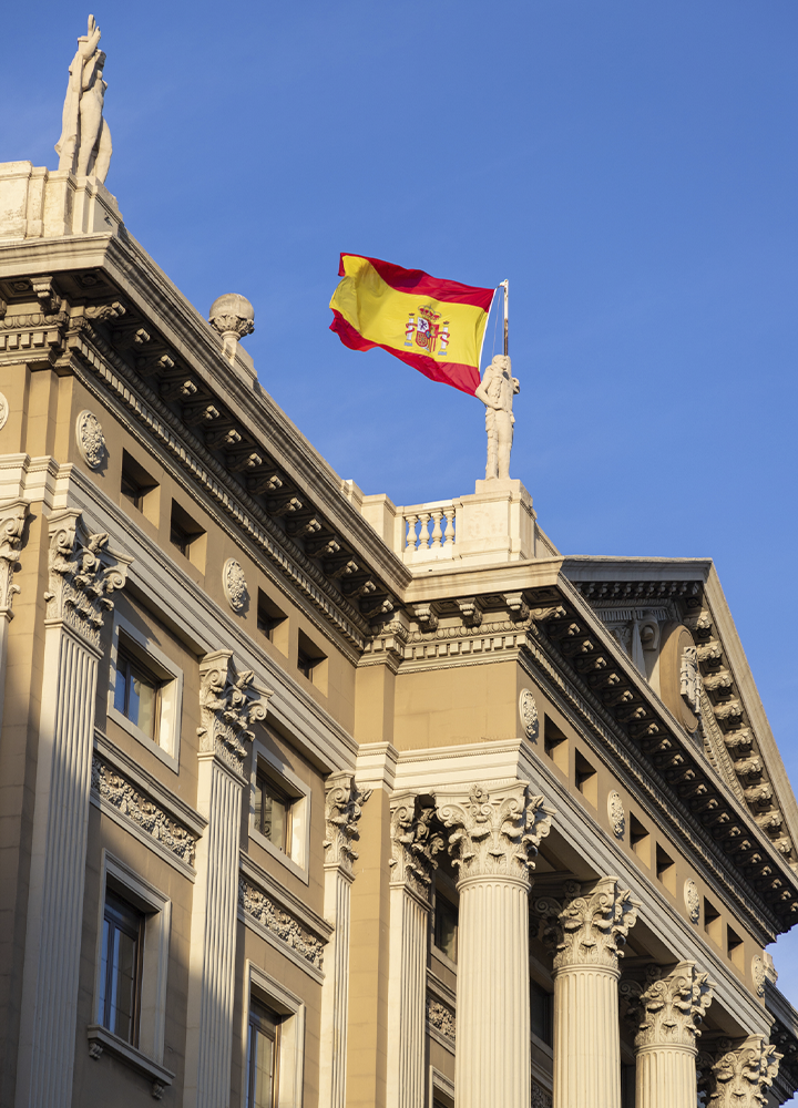 Bandera de España ondeando sobre la azotea de un edificio de arquitectura clásica, con columnas ornamentadas y esculturas en la parte superior. El cielo azul despejado ocupa gran parte del fondo y realza los tonos cálidos de la fachada.