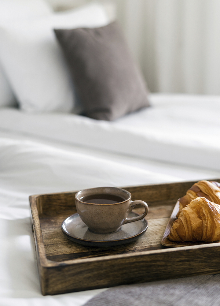 Bandeja de madera sobre una cama con ropa de cama blanca. En la bandeja hay una taza de café sobre un plato y dos cruasanes dorados. Al fondo, desenfocados, se distinguen una almohada blanca y un cojín gris, creando un ambiente de alojamiento cómodo y tranquilo.