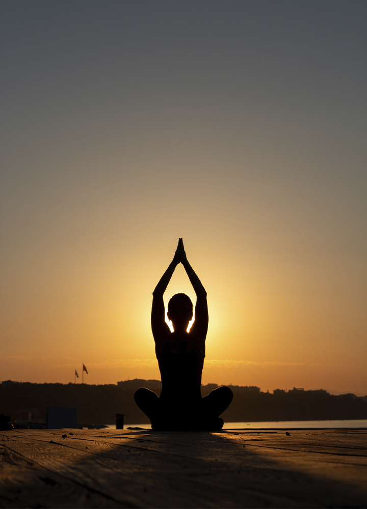 Silueta de una persona sentada en posición de meditación sobre una plataforma al aire libre, con las manos unidas sobre la cabeza. El sol se encuentra justo detrás, iluminando el cielo con tonos cálidos de atardecer.