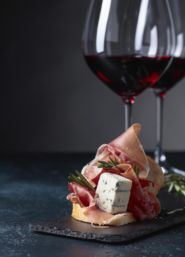 Aperitivo gourmet servido sobre una base de pan y tabla de pizarra, con lonchas de jamón curado, queso azul y ramitas de romero. Al fondo aparecen dos copas de vino tinto desenfocadas sobre una superficie oscura, en una composición elegante con iluminación tenue que resalta los colores y texturas de los alimentos.