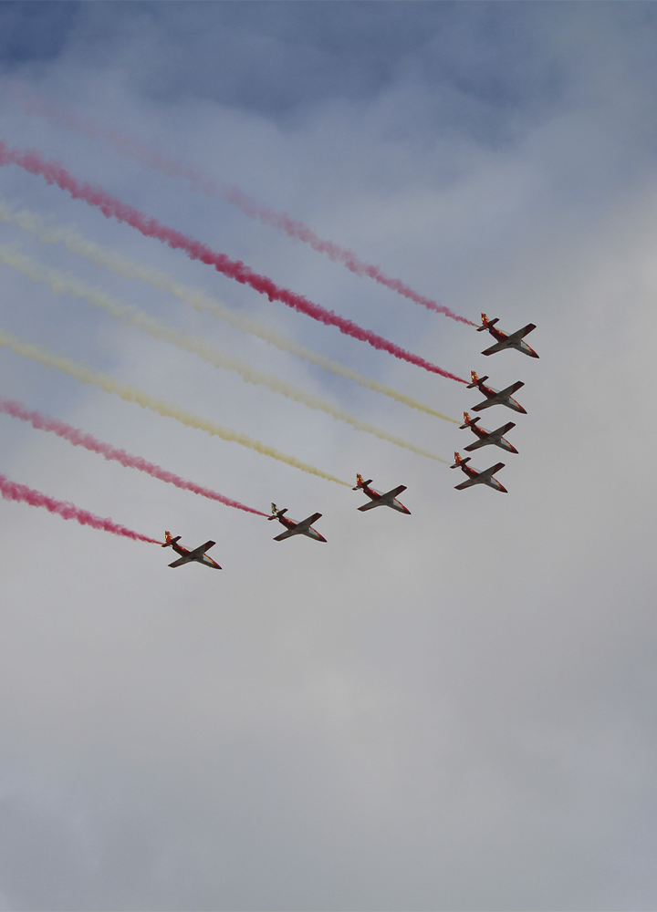 Siete aviones vuelan en formación sobre un cielo nublado, dejando estelas de humo rojo y amarillo que dibujan líneas diagonales a su paso.