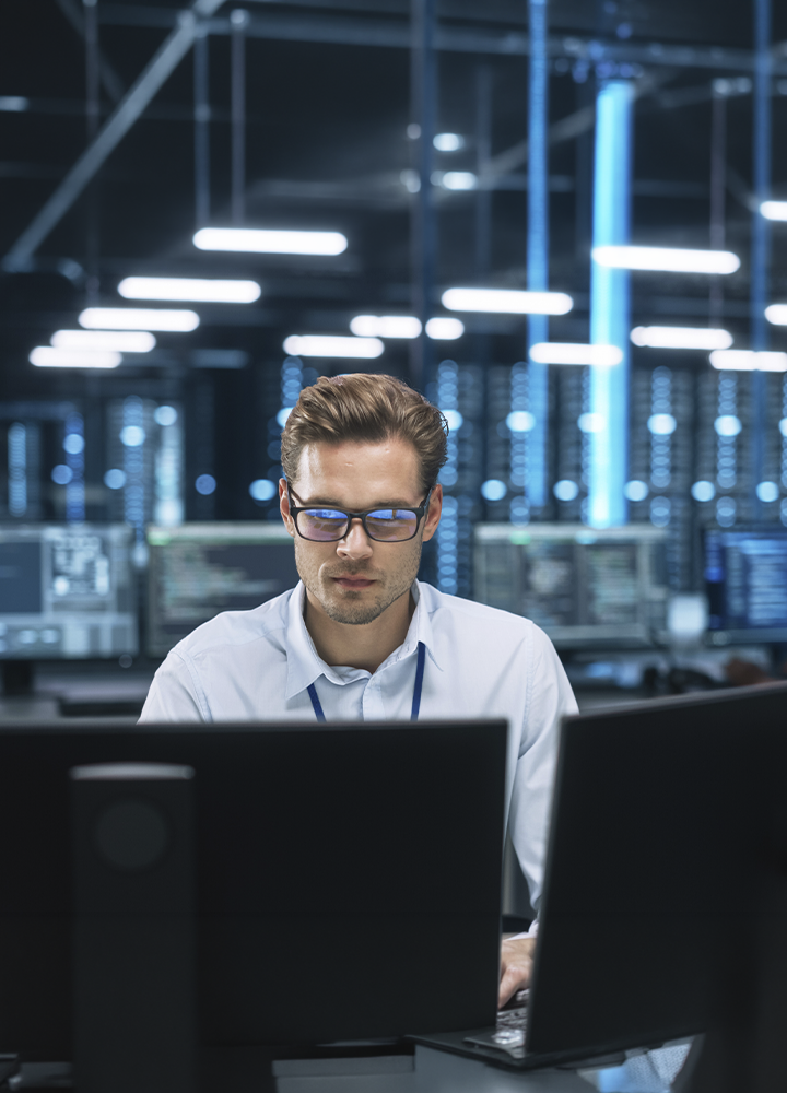 Persona trabajando frente a varios monitores en una oficina tecnológica, con pantallas y equipos informáticos alrededor. La iluminación azulada y el entorno moderno refuerzan la sensación de trabajo digital y análisis de datos.