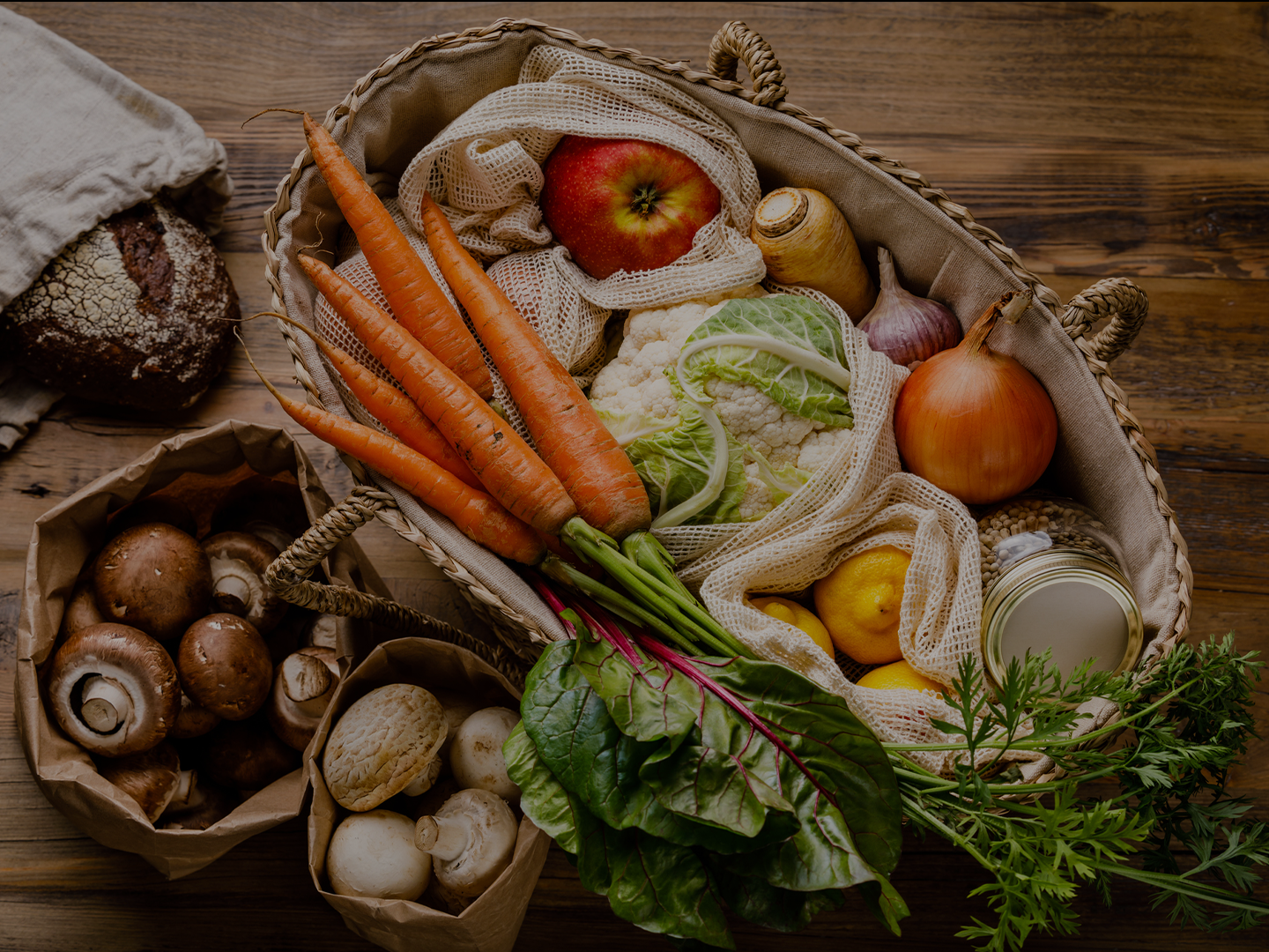 Cesta de mimbre con frutas, verduras y productos frescos en bolsas reutilizables sobre una mesa de madera. Incluye zanahorias, coliflor, cebolla, limón, manzana, champiñones y pan, asociada a una compra sostenible y de proximidad.