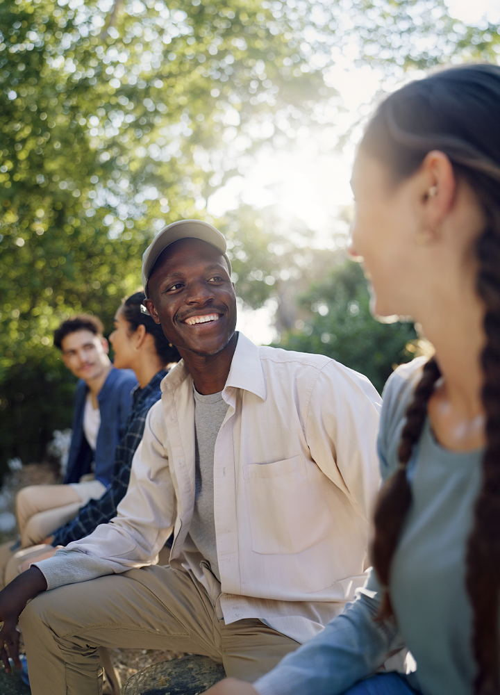 Grupo de personas sentadas al aire libre en un entorno rodeado de árboles, conversando en un ambiente relajado. En primer plano destaca una persona con gorra clara y camisa beige mirando hacia otra persona situada a la derecha, mientras al fondo se distinguen más participantes desenfocados entre la vegetación iluminada por la luz del sol.