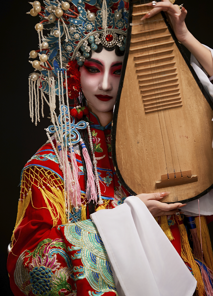 Persona con vestimenta tradicional de ópera china sosteniendo un instrumento de cuerda de madera. Destacan el elaborado tocado adornado con perlas y bordados de colores intensos sobre un fondo negro.