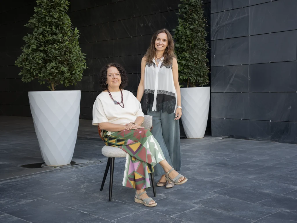 Dos personas posan en un espacio exterior de diseño contemporáneo. Una está sentada en una silla tapizada y la otra permanece de pie a su lado, frente a una fachada oscura y flanqueadas por grandes maceteros blancos con arbustos.