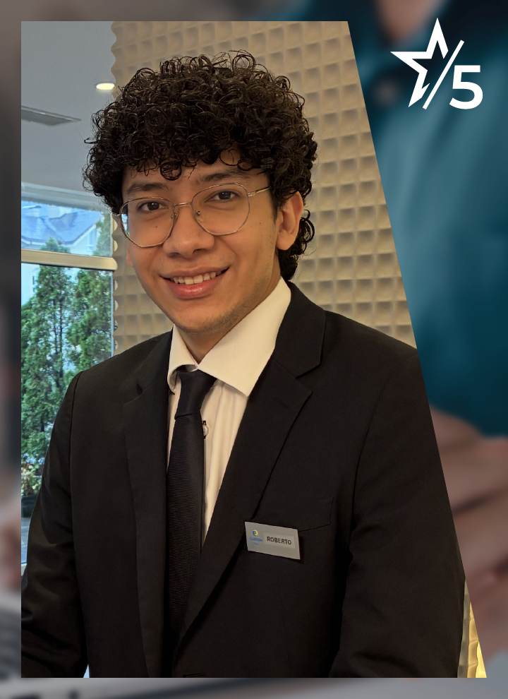 Persona con traje oscuro, camisa blanca, corbata y gafas posa en un espacio interior luminoso. Lleva una placa identificativa en la chaqueta y, en la esquina superior derecha, aparece el símbolo de una estrella junto al número 5.