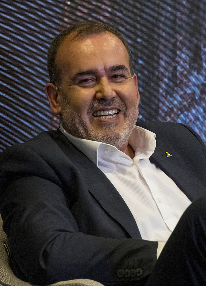 Persona sentada en un sillón durante un evento o conferencia, vestida con americana oscura y camisa blanca. El retrato está realizado en primer plano, con una pantalla iluminada como fondo y una insignia corporativa visible en la solapa.