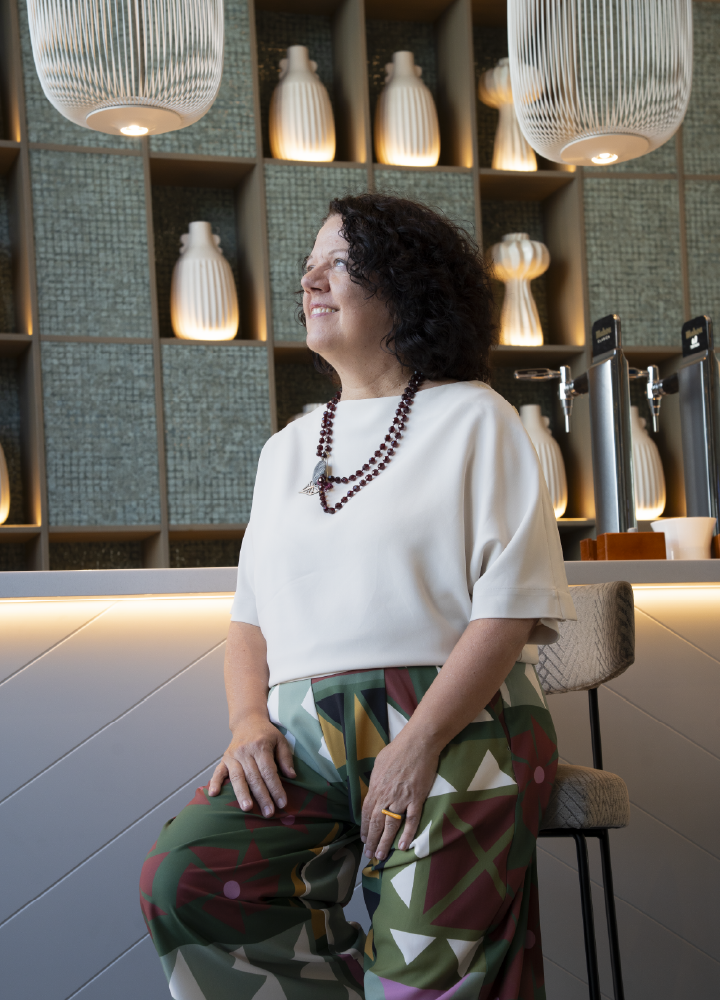 Persona sentada en un asiento tapizado dentro de un espacio interior moderno. Viste una blusa clara de manga corta, un collar de cuentas en dos vueltas y un pantalón de estampado geométrico multicolor. Al fondo hay estanterías con piezas decorativas blancas iluminadas con luz cálida.