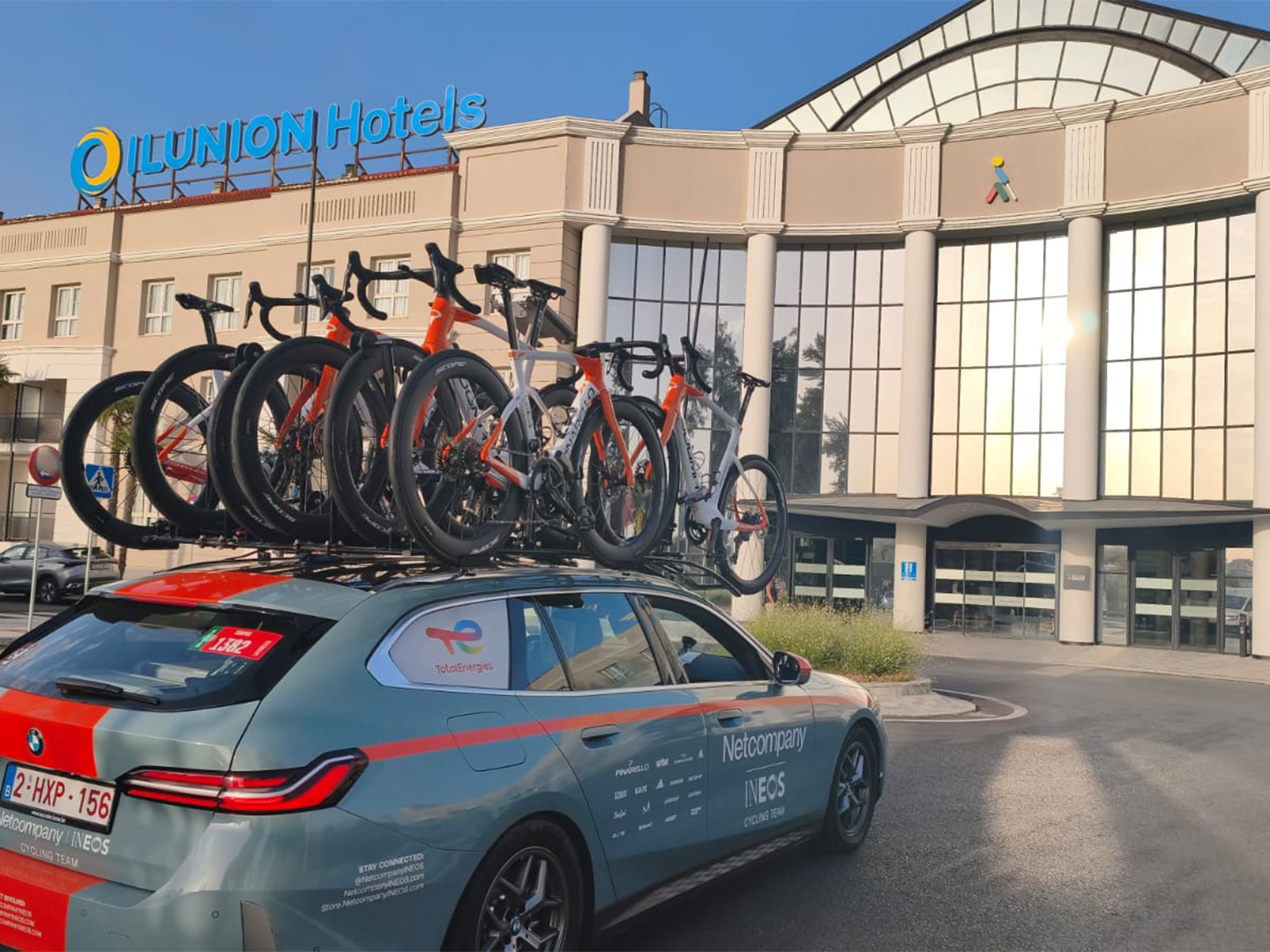 Vehículo de apoyo del equipo ciclista INEOS con varias bicicletas de competición sobre el techo, estacionado frente a la entrada principal de un hotel ILUNION. La fachada del edificio destaca por sus grandes ventanales y columnas, con el logotipo de ILUNION Hotels visible en la parte superior.