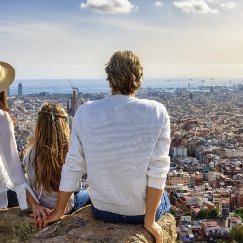 Tres personas sentadas sobre una roca contemplan una amplia vista panorámica de una ciudad costera desde un mirador elevado. Al fondo se distinguen numerosos edificios, el mar en el horizonte y varias embarcaciones, bajo un cielo despejado con algunas nubes dispersas.
