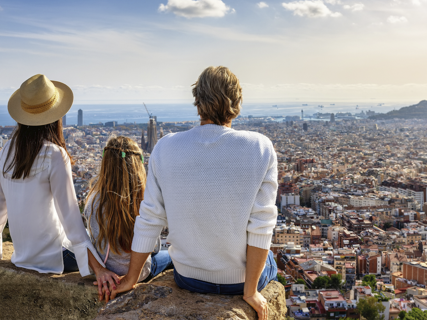 Tres personas sentadas sobre una roca contemplan una amplia vista panorámica de una ciudad costera desde un mirador elevado. Al fondo se distinguen numerosos edificios, el mar en el horizonte y varias embarcaciones, bajo un cielo despejado con algunas nubes dispersas.