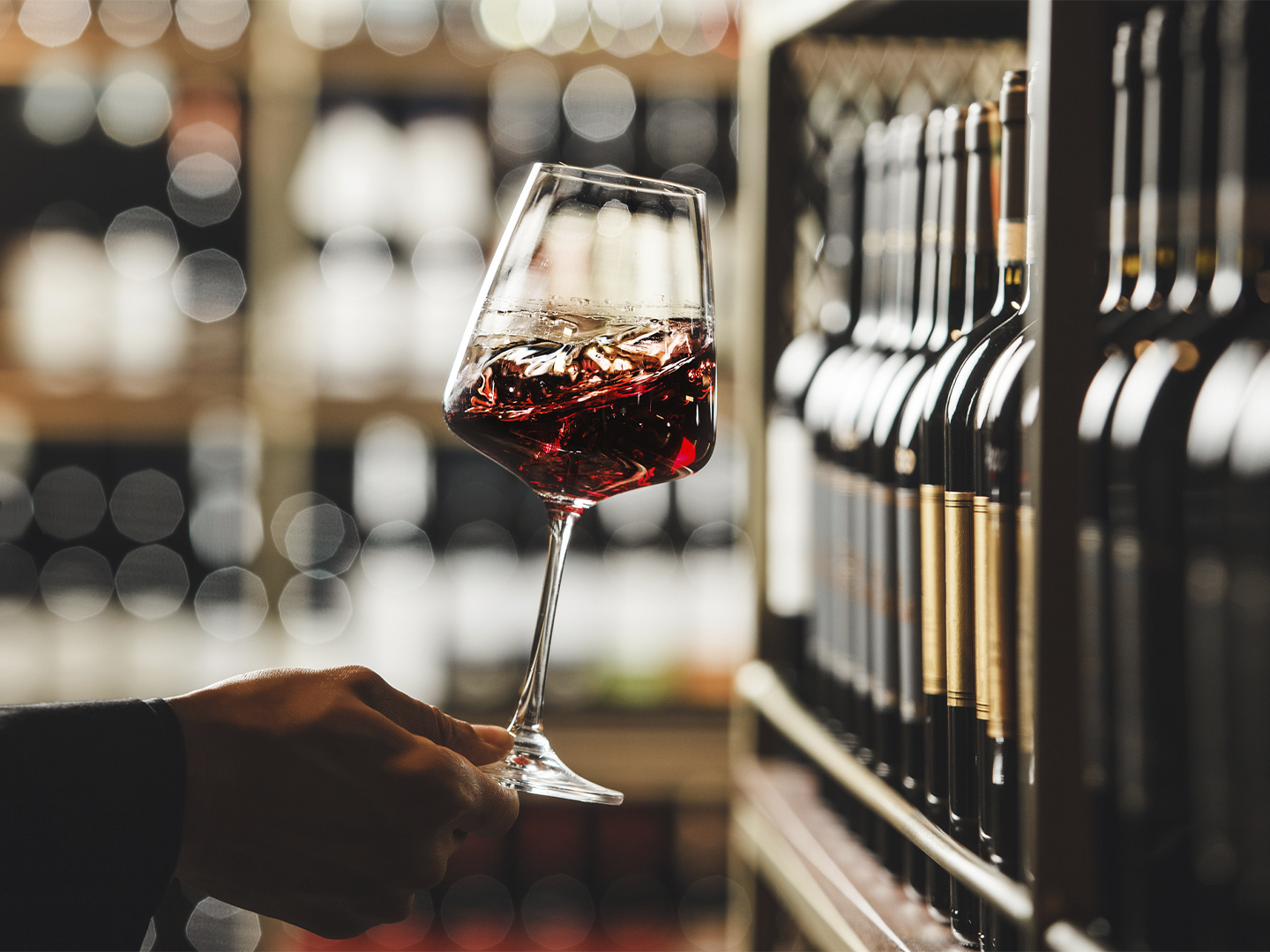 Copa de vino tinto sostenida por una mano frente a una estantería con botellas alineadas. El vino aparece en movimiento dentro de la copa, destacando los reflejos y tonalidades rojizas. El fondo desenfocado muestra una bodega o tienda especializada, con iluminación cálida y puntos de luz que realzan el ambiente.