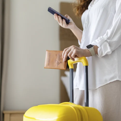 Persona en el interior de una habitación sosteniendo un teléfono móvil junto a una maleta amarilla con asa extensible. Una mano descansa sobre el asa mientras sostiene un pasaporte marrón, y en la muñeca lleva un reloj inteligente. El entorno está iluminado por luz natural y sugiere una situación de preparación o gestión de un viaje.