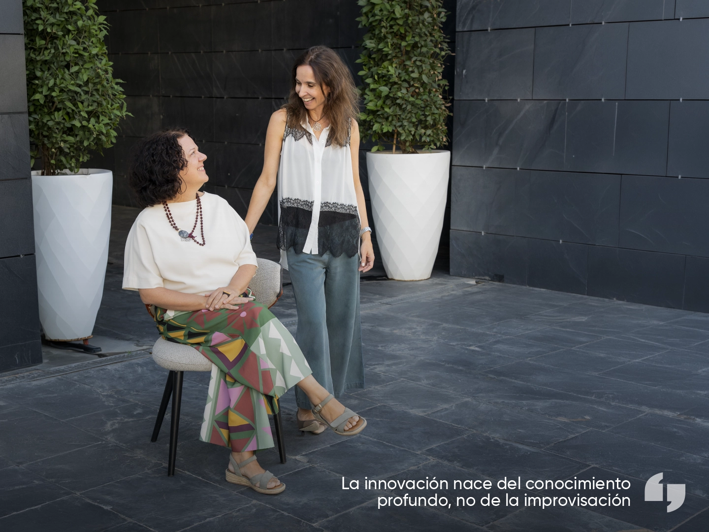 Dos personas interactúan en un espacio exterior con suelo de piedra gris y una pared revestida con paneles oscuros. Una persona está sentada en una silla tapizada y la otra permanece de pie a su lado. A ambos lados de la escena hay grandes macetas blancas con arbustos verdes. En la esquina inferior derecha aparece el texto: “La innovación nace del conocimiento profundo, no de la improvisación”.