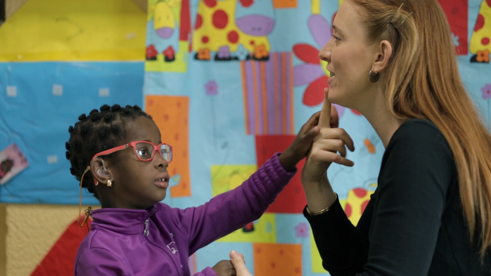 Persona adulta y una niña interactúan en un entorno educativo, mientras la niña toca suavemente la barbilla de la persona adulta. Al fondo se ven murales coloridos y materiales decorativos propios de un aula infantil.