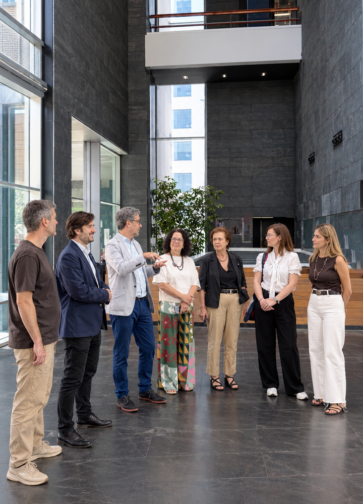 Grupo de siete personas conversando en el vestíbulo de un edificio moderno de techos altos y grandes ventanales. El espacio cuenta con paredes oscuras, suelo de piedra y abundante luz natural. Una de las personas interviene mientras el resto permanece reunido en semicírculo, en un entorno de encuentro profesional.