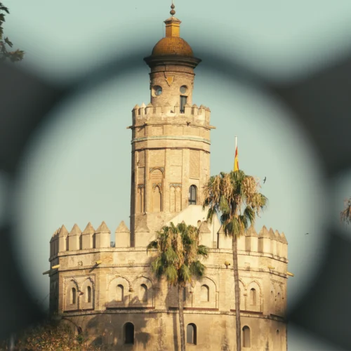 Vista de la Torre del Oro de Sevilla encuadrada a través de una estructura desenfocada en primer plano que crea una abertura circular alrededor del monumento. La torre de piedra, coronada por una cúpula dorada, se alza entre palmeras y vegetación, con una bandera española visible junto a uno de sus laterales. El cielo claro y la luz cálida resaltan los detalles arquitectónicos de este emblemático edificio histórico.
