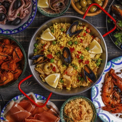 Vista cenital de una mesa de madera con una paella de marisco en el centro, decorada con mejillones, gambas, guisantes y gajos de limón. Alrededor se distribuyen varios platos de cocina mediterránea, entre ellos calamares con aceitunas, mejillones, jamón curado, hummus, patatas especiadas, gambas a la plancha y aceitunas.