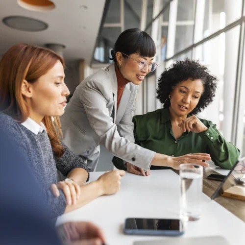 Tres personas reunidas alrededor de una mesa de trabajo observan una tableta colocada sobre un soporte junto a una gran ventana. Una de las personas señala la pantalla mientras las demás siguen la explicación. Sobre la mesa hay un vaso de agua, un teléfono móvil y unas gafas, en un espacio de oficina iluminado por luz natural.