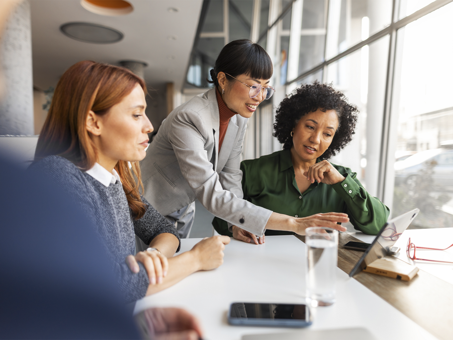 Tres personas reunidas alrededor de una mesa de trabajo observan una tableta colocada sobre un soporte junto a una gran ventana. Una de las personas señala la pantalla mientras las demás siguen la explicación. Sobre la mesa hay un vaso de agua, un teléfono móvil y unas gafas, en un espacio de oficina iluminado por luz natural.