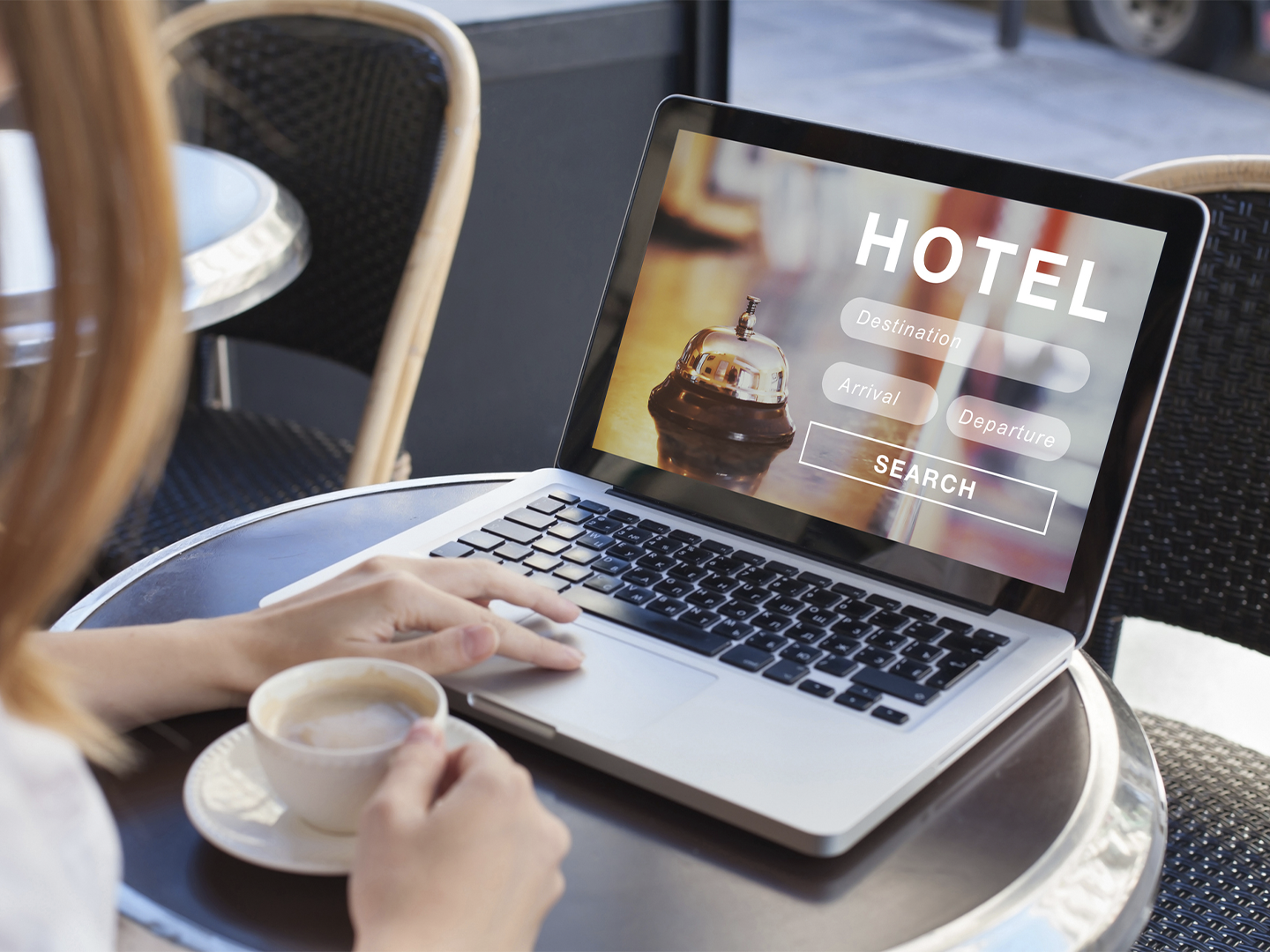 Persona sentada en una mesa de cafetería utilizando un ordenador portátil con una página de búsqueda de hoteles abierta. En la pantalla aparece el texto “HOTEL”, junto a campos para destino, llegada y salida, y un botón de búsqueda. Sobre la mesa hay una taza de café en primer plano y una mano apoyada en el teclado. El entorno cuenta con sillas de mimbre oscuro y luz natural procedente de un ventanal cercano.
