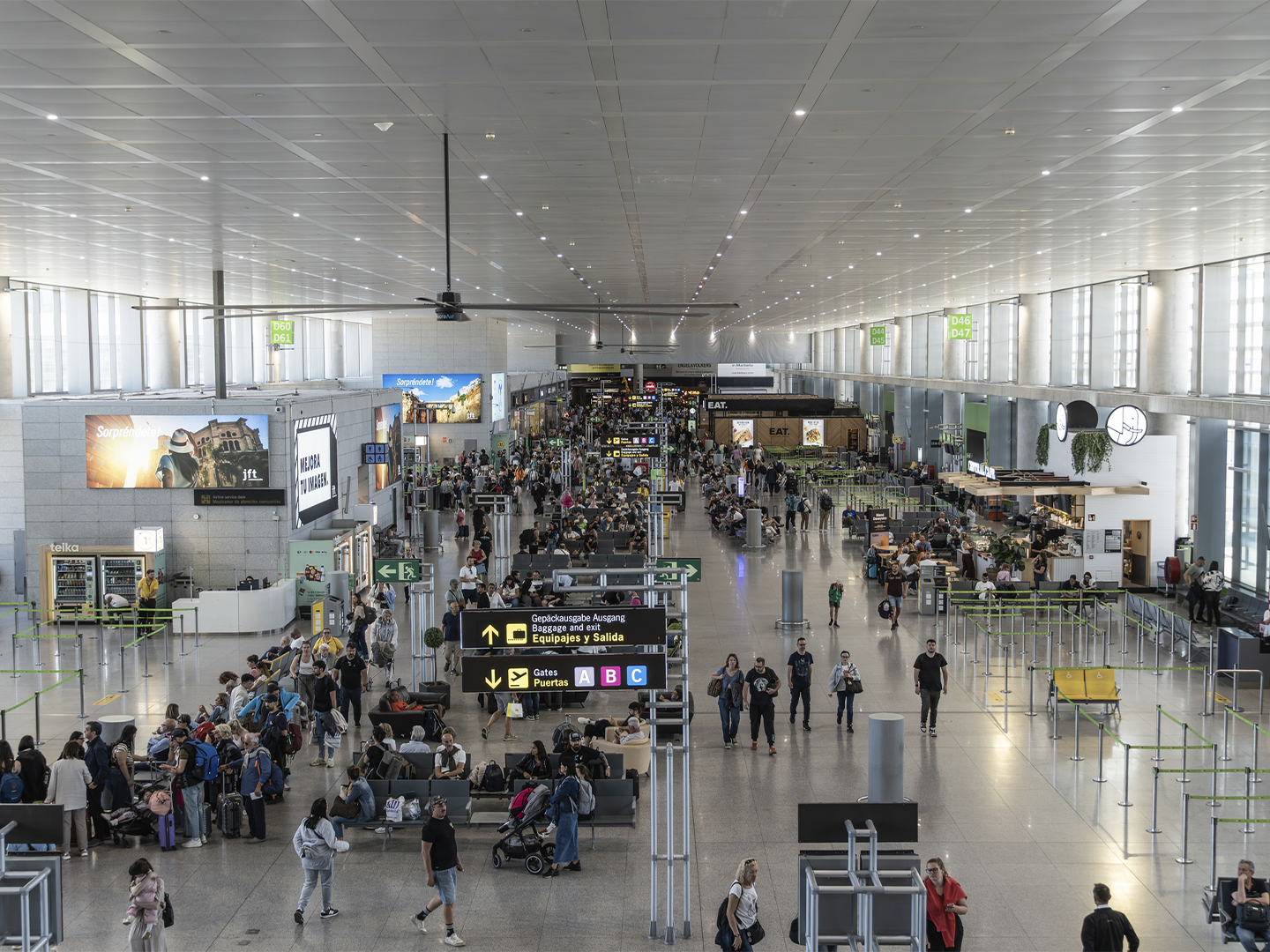 Amplia terminal de aeropuerto con numerosos viajeros, zonas de espera y controles de seguridad. En el centro destacan paneles de señalización con indicaciones hacia puertas de embarque y recogida de equipajes, mientras tiendas y áreas de restauración se distribuyen a ambos lados del recinto.